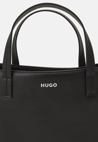Crna kožna torba sa dva gornja ručka, sa zrnatim teksturom i srebrnim "HUGO" logom na prednjoj strani. Klasično strukturiran dizajn.