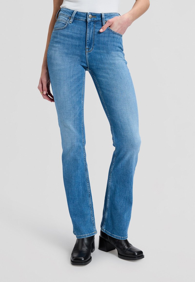 Femme portant un jean droit bleu clair et des bottines noires, debout avec une main dans la poche devant un fond uni.