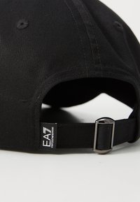 Boné de beisebol em algodão preto com textura em costura, faixa ajustável com fivela metálica e um rótulo de marca na lateral.