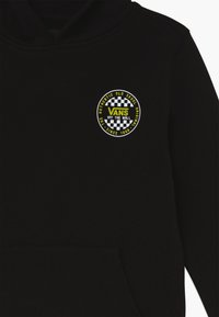 Sudadera negra con capucha que presenta un logo bordado circular en amarillo y blanco con un borde a cuadros. Tela suave, bolsillo tipo canguro en la parte delantera.