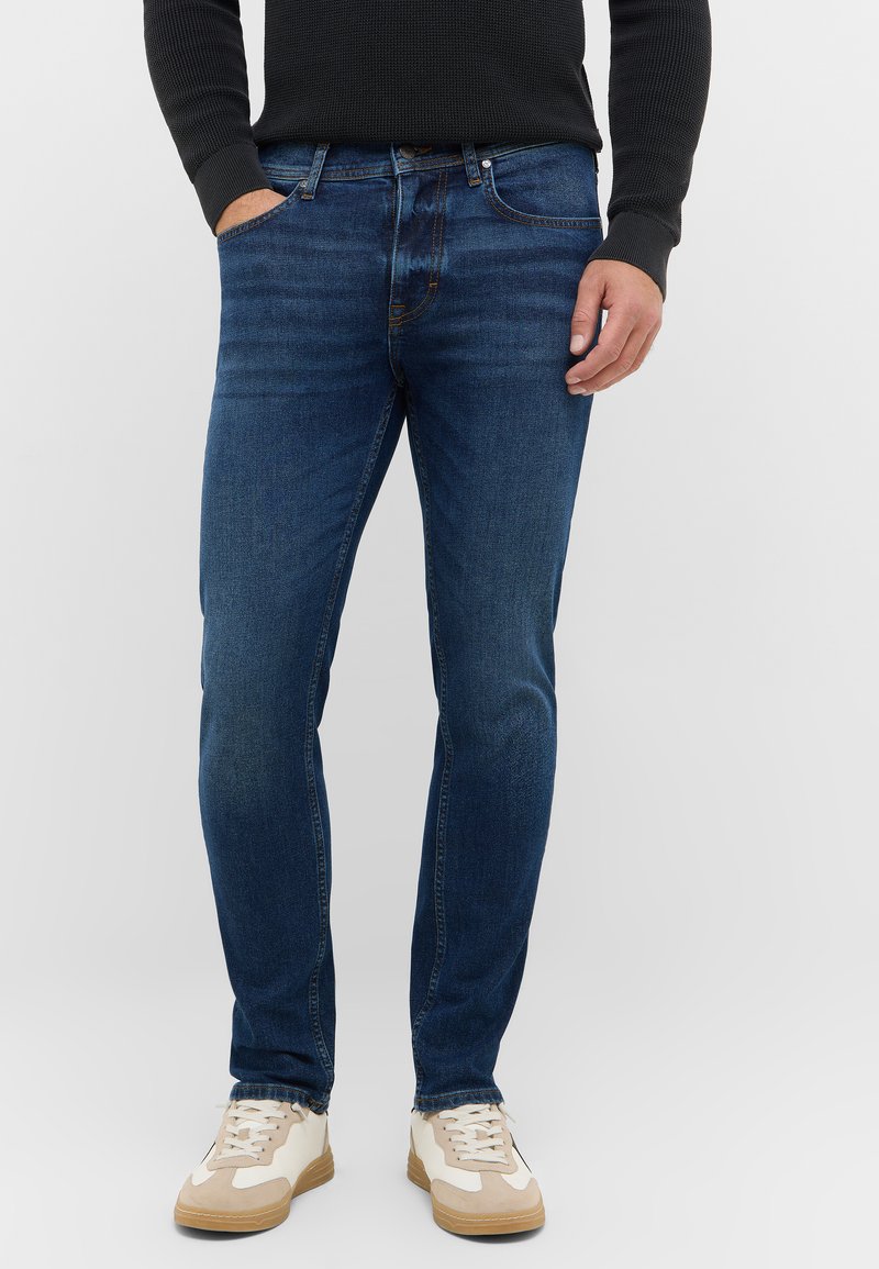 Granatowe dżinsy o slim fit, z pięcioma kieszeniami i subtelnym wyblaknięciem. Noszone z jasnymi butami sportowymi.