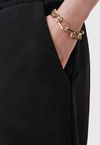 Zwarte katoenen sweatpants met een zak en een reliëflogo. Een gouden kettingarmband is zichtbaar om de pols.
