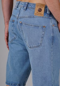 Light blue denim shorts with a relaxed fit, featuring back pockets, copper rivets, and a branded leather patch at the waistband.
