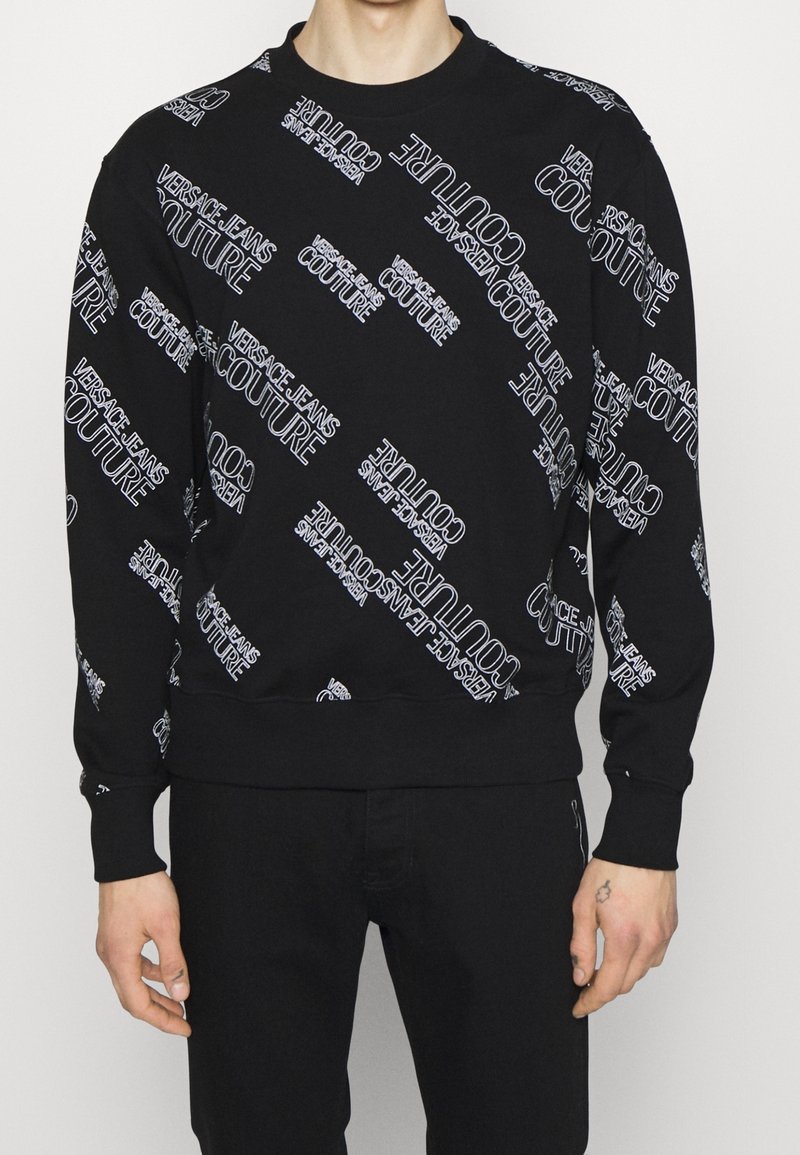 Sweat-shirt noir à manches longues avec texte blanc « Versace Jeans Couture » imprimé en diagonale sur le devant et les manches.