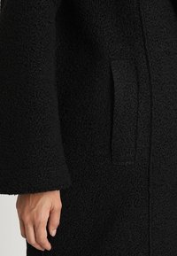 Černý, strukturovaný kabát s jemným povrchem, obsahující boční kapsu a široké rukávy. Materiál se jeví jako teplý a silný.