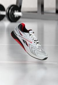 Άσπρο, γκρι και κόκκινο αθλητικό παπούτσι Reebok με μαύρες λεπτομέρειες, αιωρούμενο σε γυμναστήριο με θολά βάρη και εξοπλισμό στο παρασκήνιο.