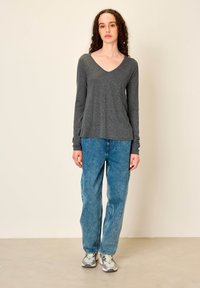 Moteris su garbanotais plaukais, vilkinti tamsiai pilką ilgomis rankovėmis V formos kaklo palaidinę, mėlynas džinsus ir pilkus sportbačius, stovinti priešais vienodus smėlio spalvos foną.