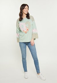 Mentazöld felső, szív mintával textil foltokkal, kockás ujjak és virágminták díszítik. Kék skinny farmernadrággal és fehér cipővel viselt.