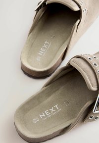 Beige velúr papucs, sima felülettel, állítható pánttal, ezüst színű kiegészítőkkel és párnázott talppal, amelyen a márka logója található.
