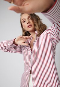 Kobieta w czerwono-białą poziomą w paski koszulę z guzikami, unosząca ręce, częściowo zasłaniająca obiektyw kamery.