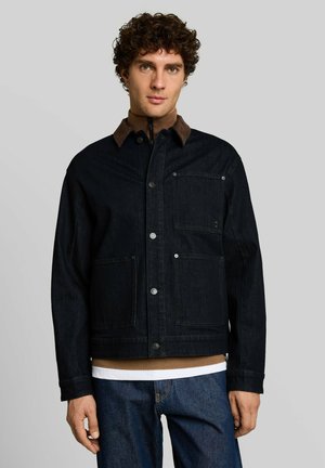 Junger Mann mit lockigem Haar, der eine dunkle Jeansjacke mit aufgesetzten Taschen über einem braunen Rollkragenpullover und blauen Jeans trägt und vor einem grauen Hintergrund steht.