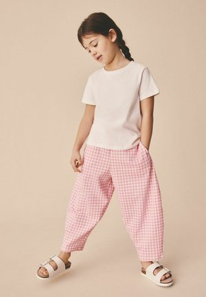 Młoda dziewczyna z warkoczami, ubrana w białą koszulkę, różowe szerokie spodnie w kratę i białe sandały na klamrę na beżowym tle.