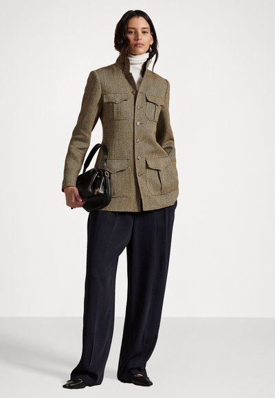 Sako z tweedu s houndstooth vzorem, s dvěma předními kapsami, v přiléhavém střihu, v hnědých odstínech, nošeno přes bílé rolák, s černou kabelkou a kalhotami.