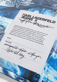 Blå mönstrad tyg med en quiltad textur, med en vit etikett som visar tryckt text och en signatur. Begränsad upplaga design detaljer.