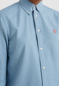 Camicia in cotone azzurro chiaro con colletto classico, bottoni bianchi e un piccolo logo ricamato in corallo sul petto.