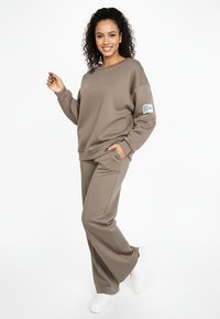 Jasnobrązowy komplet z bluzy i spodni. Bluza ma okrągły dekolt i naszywkę na lewym rękawie. Miękka, teksturowana tkanina. Białe trampki.