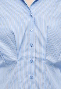 Μπλε και λευκό πουκάμισο με κάθετες ρίγες και γιακά, με πέντε μπλε κουμπιά κουμπωμένα κατά μήκος του κέντρου μπροστά.
