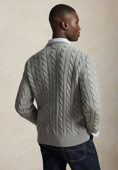 Uomo che indossa un maglione a trecce grigio chiaro sopra una camicia azzurra con colletto, abbinato a jeans scuri, di profilo con le mani nelle tasche.