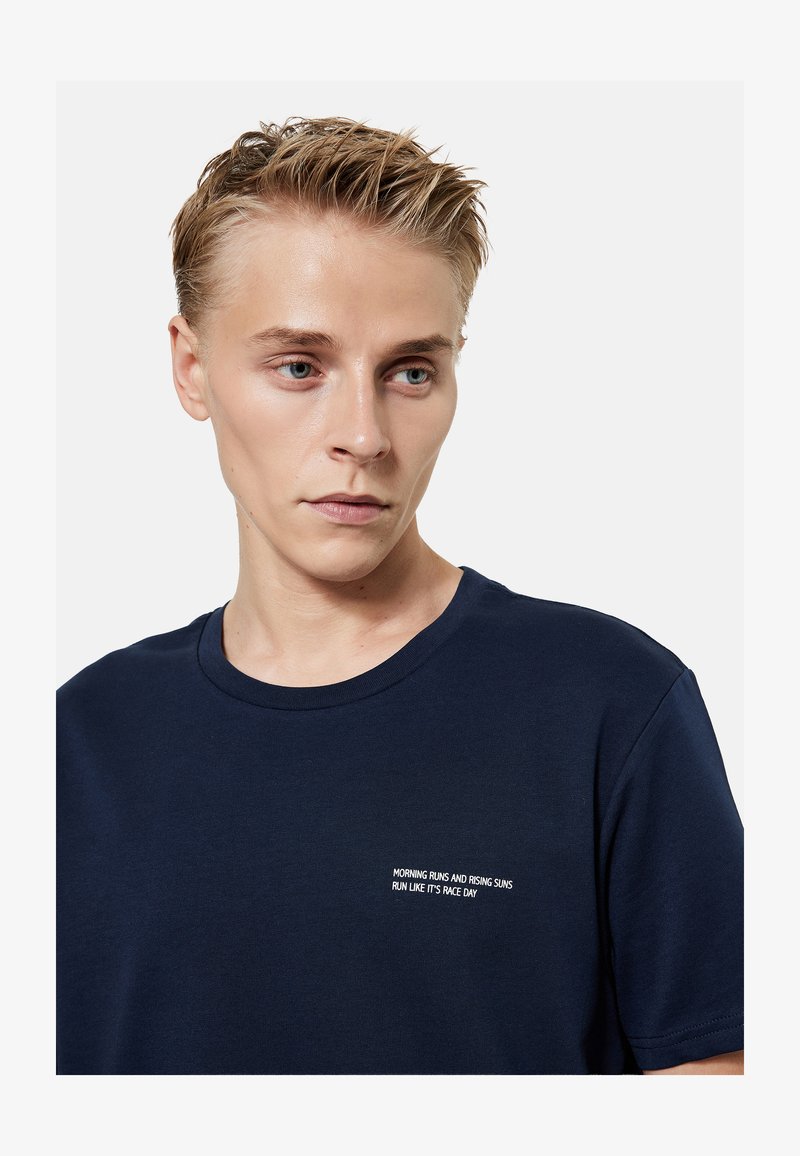 Jeune homme aux cheveux blonds courts portant un T-shirt bleu marine avec un texte blanc : « Courses matinales et soleils levant courent comme si c’était jour de course. »