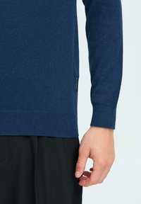Suéter de punto azul marino con manga larga y puños acanalados. Presenta un dobladillo recto con una etiqueta visible en el lado.