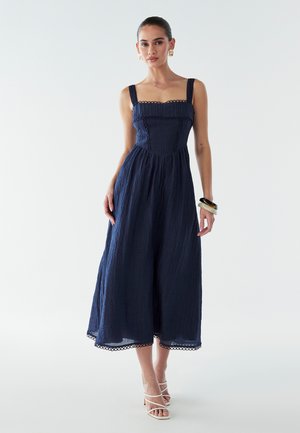 Robe midi bleu marine avec un décolleté carré, tissu texturé, bordures en dentelle et bretelles ajustables, assortie à des sandales blanches à lanières.