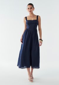 Vestido midi azul marino con escote cuadrado, tela texturizada, detalles de encaje y tirantes anchos, combinado con sandalias blancas de tiras.