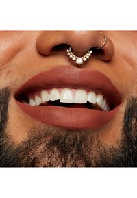 Närbild av läppar med matt rödbrun läppstift, som framhäver vita tänder, förstärkt med en dekorativ näsring med pärlor och en halvmåneform.