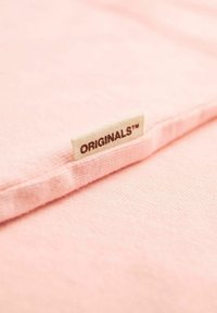 Gaiši rozā audums ar nedaudz teksturētu virsmu. Malā ir piešūta etiķete ar uzrakstu "ORIGINALS™".