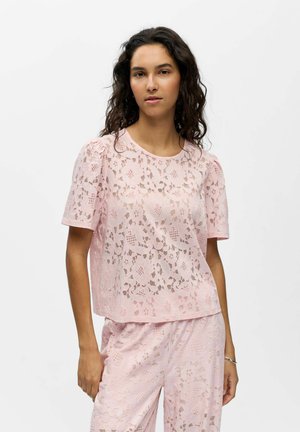 Femme portant un haut en dentelle rose clair à manches courtes et un pantalon assorti, debout avec une main dans la poche sur un fond uni.