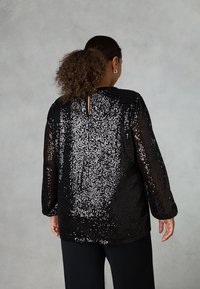 Top nero in paillettes con maniche lunghe, caratterizzato da un design con apertura a forma di chiave sulla schiena. La texture è lucida con un tessuto morbido al di sotto.