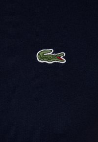 Logo de crocodile vert brodé avec une bouche rouge et un contour blanc sur fond en tissu côtelé bleu marine foncé.