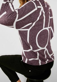 Maglione viola con motivi geometrici bianchi, caratterizzato da un tessuto strutturato e una vestibilità morbida. Indossato con pantaloni neri, seduto su uno sgabello.