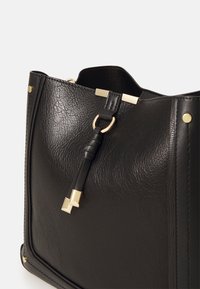 Czarna skórzana torebka z metalowymi elementami w złotym kolorze oraz skórzaną frędzlą zawieszoną na okrągłym złotym kółku w pobliżu górnej krawędzi.