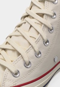 Κοντινό πλάνο αθλητικού παπουτσιού από εκρού καμβά με λευκά κορδόνια, περασμένα μέσα από ασημένιες τρύπες, και μια κόκκινη ρίγα κατά μήκος της άκρης της σόλας.