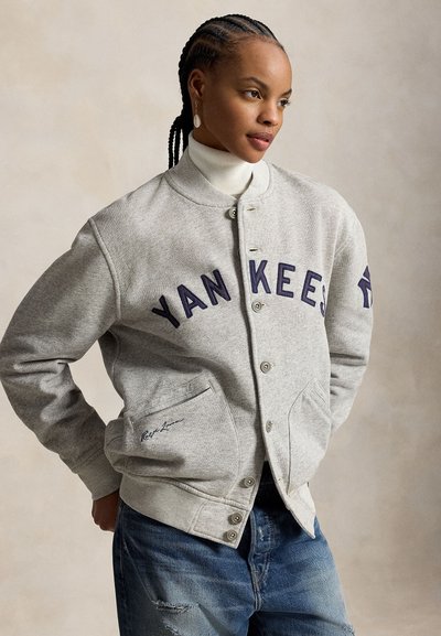 Femme aux cheveux tressés portant une veste boutonnée gris clair avec l'inscription « YANKEES » et un jean bleu, debout les mains dans les poches.