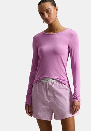 Kobieta w dopasowanym, różowym topie z długim rękawem oraz różowo-białych, paskowanych szortach z widocznym paskiem Polo Ralph Lauren.