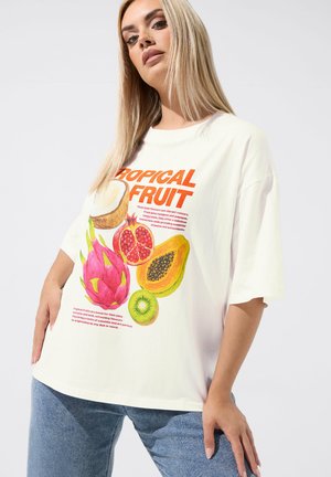 Blondynka w luźnej białej koszulce z kolorowymi tropikalnymi ilustracjami owoców i napisem, w połączeniu z niebieskimi dżinsami, na jednolitym tle.