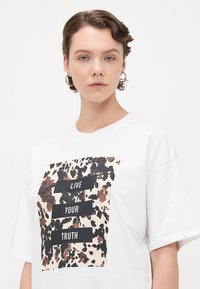 Ung person med kort brunt hår som bär en vit t-shirt med ett brunt och svart djurmönstrat tygstycke som säger "LEVA DIN SANNING."