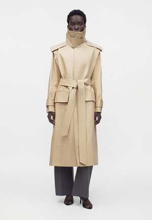 Personne portant un trench-coat beige ceinturé avec col montant, pantalon gris à jambe large et talons ouverts noirs, debout devant un fond clair uni.