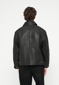 Veste en cuir noir à coupe décontractée, col large et surpiqûres visibles. Comprend des boutons décoratifs aux poignets et au dos. Texture lisse.
