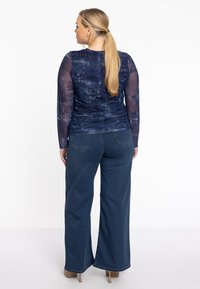 Langärmliges marineblaues Oberteil mit einem strukturierten Denim-Muster, kombiniert mit weit geschnittenen, dunkeltürkisen Jeans. Die Rückansicht zeigt eine glatte Silhouette und subtile Details.