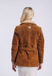 Veste en daim marron avec une taille ceinturée, un col en fausse fourrure contrastant et des manches zippées. Affiche un patch de marque dans le dos.