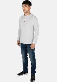 Langärmliges graues Henley-Shirt mit Rundhalsausschnitt und einer Knopfleiste mit drei Knöpfen, kombiniert mit blauen Jeans und schwarzen Sportschuhen.