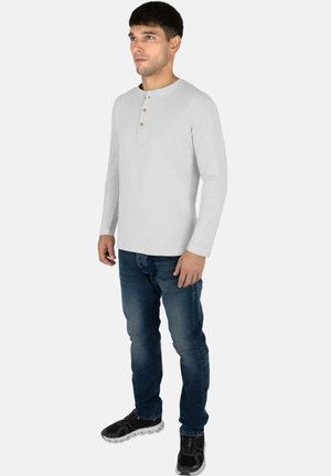 Langärmliges graues Henley-Shirt mit Rundhalsausschnitt und einer Knopfleiste mit drei Knöpfen, kombiniert mit blauen Jeans und schwarzen Sportschuhen.