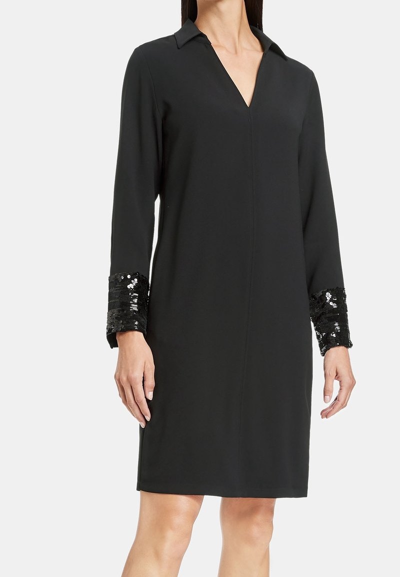 Robe noire à manches longues avec col en V et poignets noirs brillants à sequins, portée par une femme debout sur un fond clair uni.