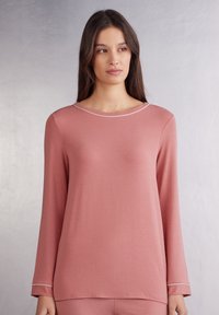 Langemanige top van zacht rozenkleurig materiaal met een ronde halslijn en subtiele witte accenten langs de kraag en mouwen. Soepele textuur, ontspannen pasvorm.