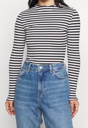 Vrouw draagt een zwart-wit horizontaal gestreept shirt met lange mouwen, ingestopt in een high-waisted blauwe denim jeans, staat met ontspannen handen.