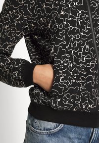 Svart mönstrad bomberjacka med abstrakta vita linjer, ribbade ärmslut och dragkedja, tillsammans med denimjeans.