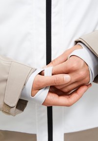 Weiße Auß Jacke mit schwarzem Reißverschluss, beigen Ärmelbündchen und grauem Innenfutter. Die Bündchen haben ein tailliertes Design mit einem Klettverschlussdetail.