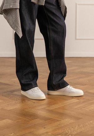 Benen in donkere wijde jeans en witte vetersneakers, staand op een lichte houten parketvloer binnen.